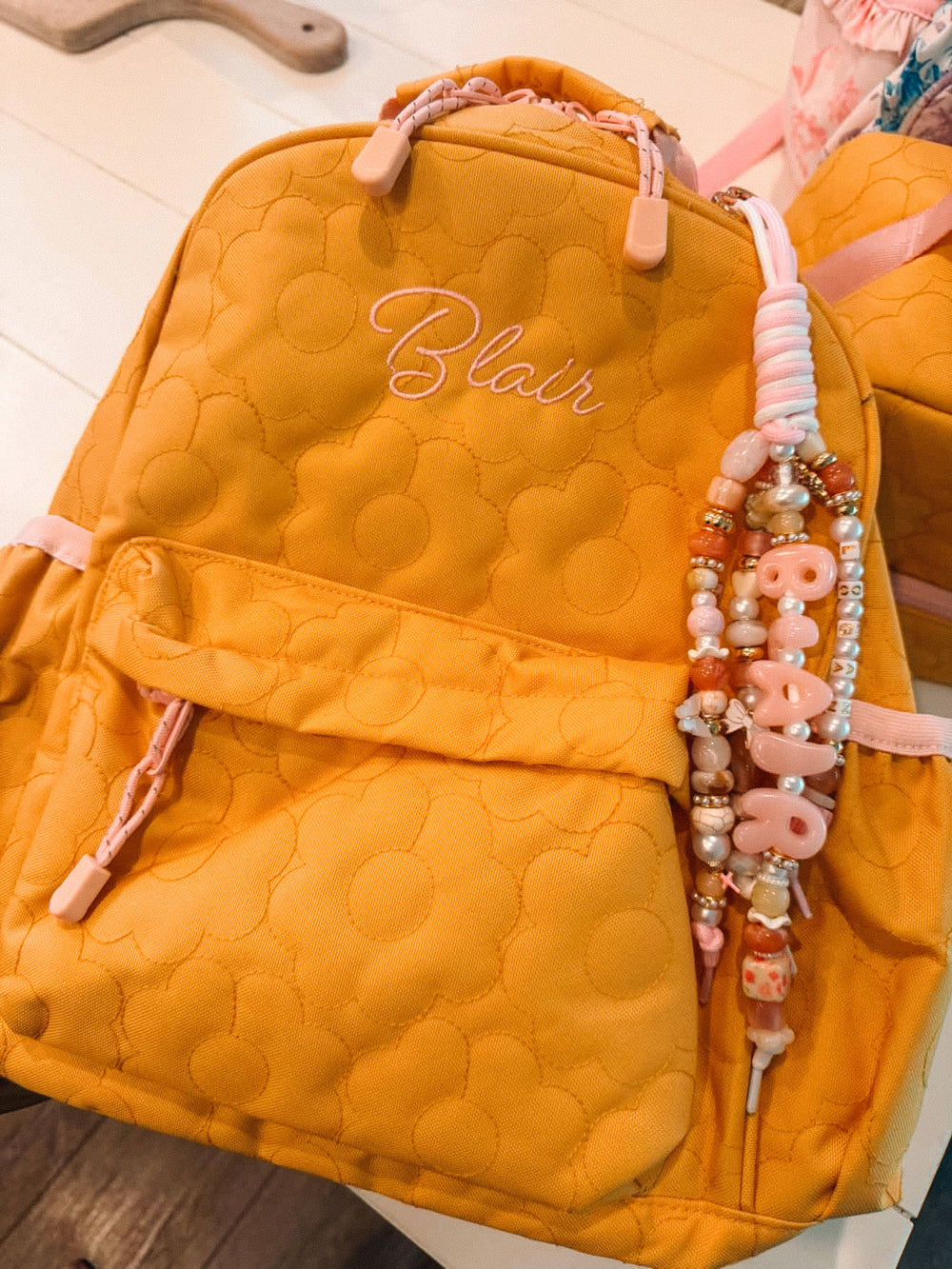 A pink and cream Lucky Loop charm spelling Blair clipped to a yellow flower-quilted backpack