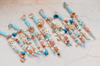 Seven personalized bag charms in blue, cream and coral laid side by side, each with a name in letter beads