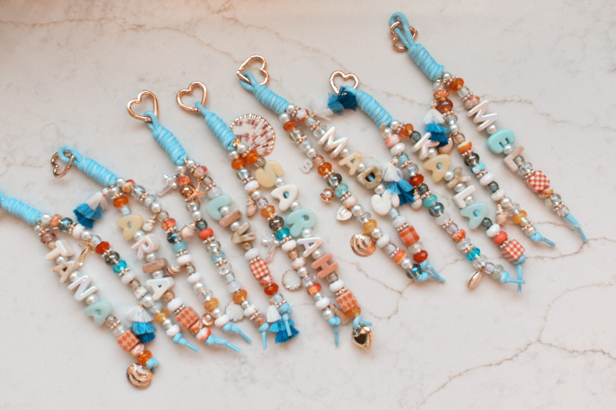 Seven personalized bag charms in blue, cream and coral laid side by side, each with a name in letter beads