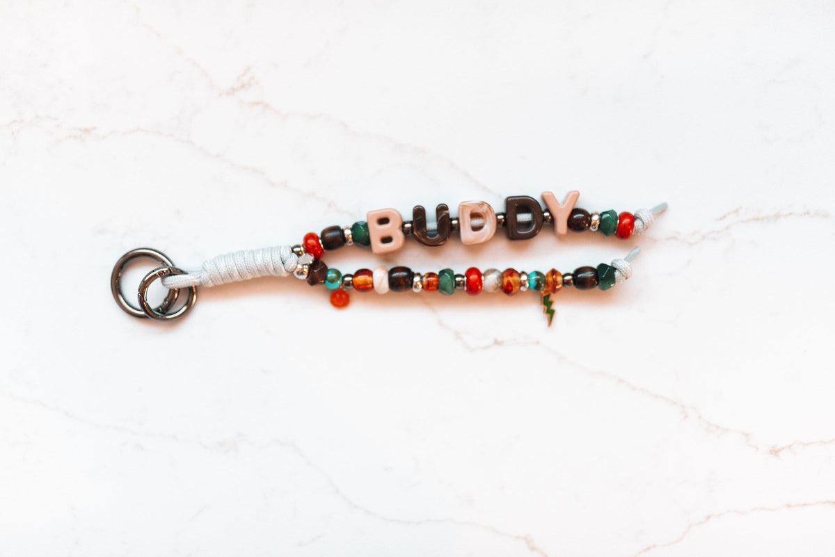 Buddy 2-strand loop with blush and chocolate BUDDY letters, earthy wood, amber, red and green beads, a lightning bolt charm and a gunmetal clasp