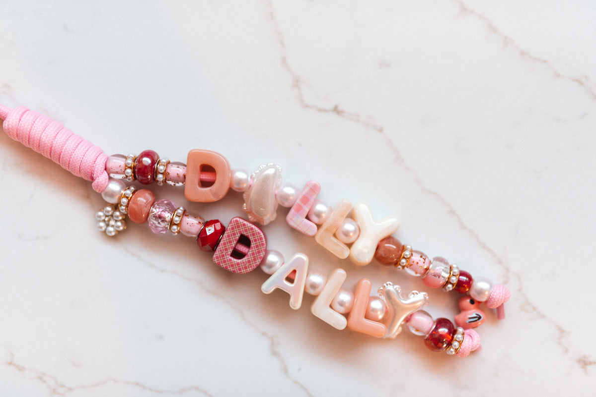 Dilly Dally 2-strand loop in shades of pink with DILLY and DALLY letter beads in blush, plaid and pearl, a pearl flower charm and a pink wrapped cord